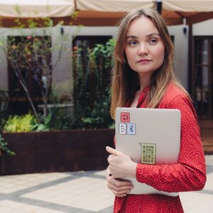 A young woman in a red dress holding a laptop outdoors, looking thoughtful.