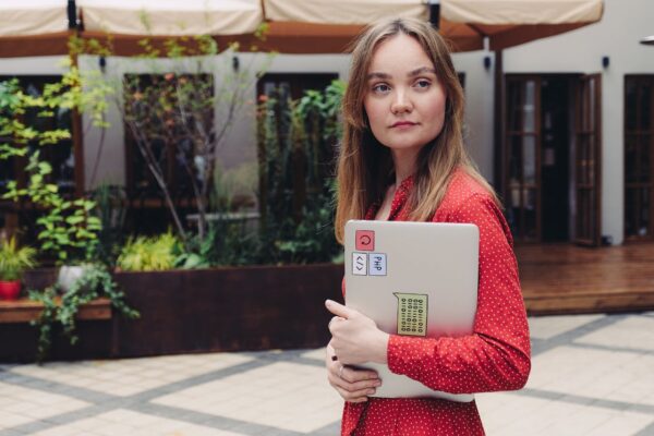 A young woman in a red dress holding a laptop outdoors, looking thoughtful.