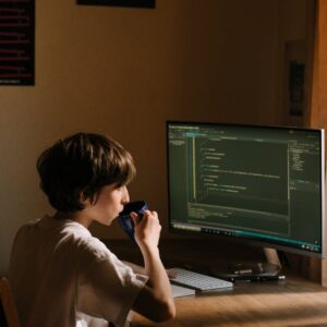 A young person coding at a desk with a computer and drinking from a mug.